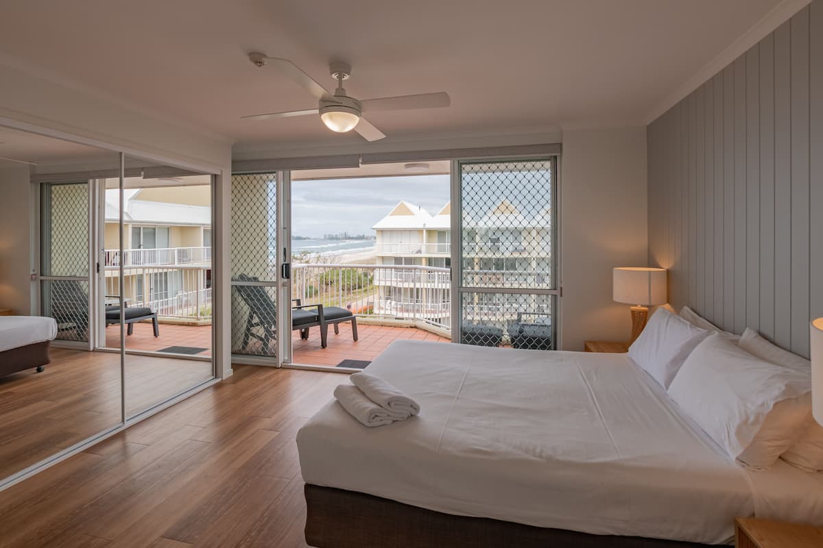 Ein Hotelzimmer mit Bett, Deckenventilator, Balkon mit Blick auf Strand und Gebäude sowie einer Schiebetür aus Glas.