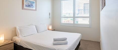 A bedroom with a bed, a bedside lamp, a ceiling fan, a window with blinds, and a framed picture on the wall.