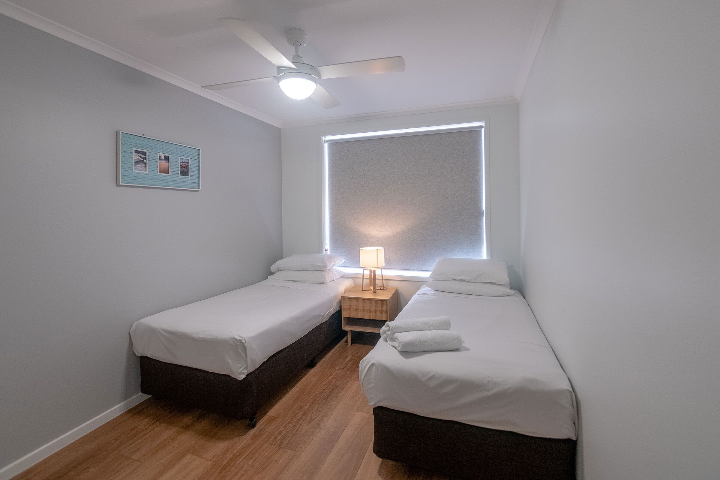 Een kamer met twee eenpersoonsbedden, een plafondventilator, een nachtkastje met een lamp en een groot raam met zonwering.