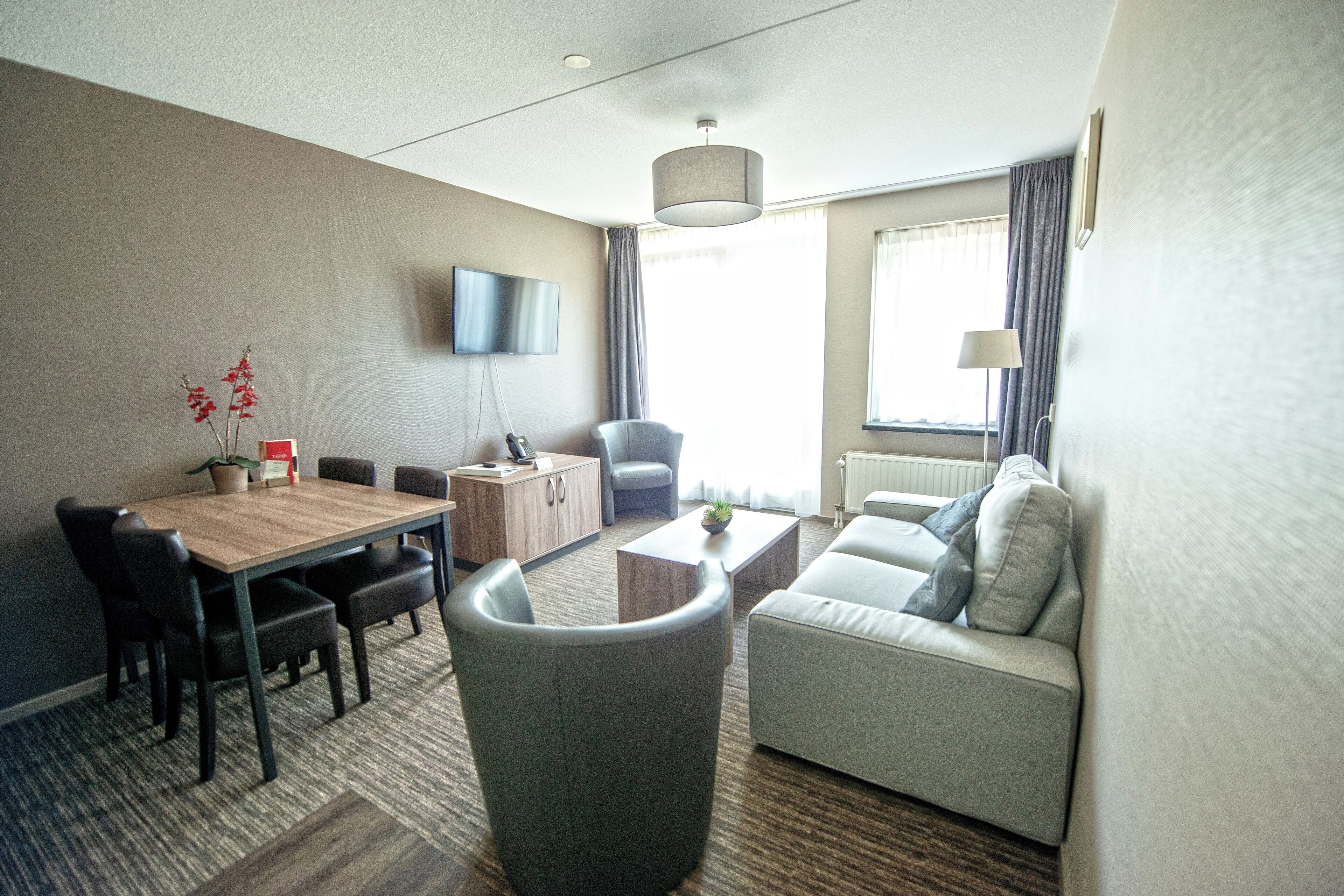 Ein modernes Hotelzimmer mit Essbereich, einer Couch, einem Fernseher und einem Fenster mit Vorhängen.