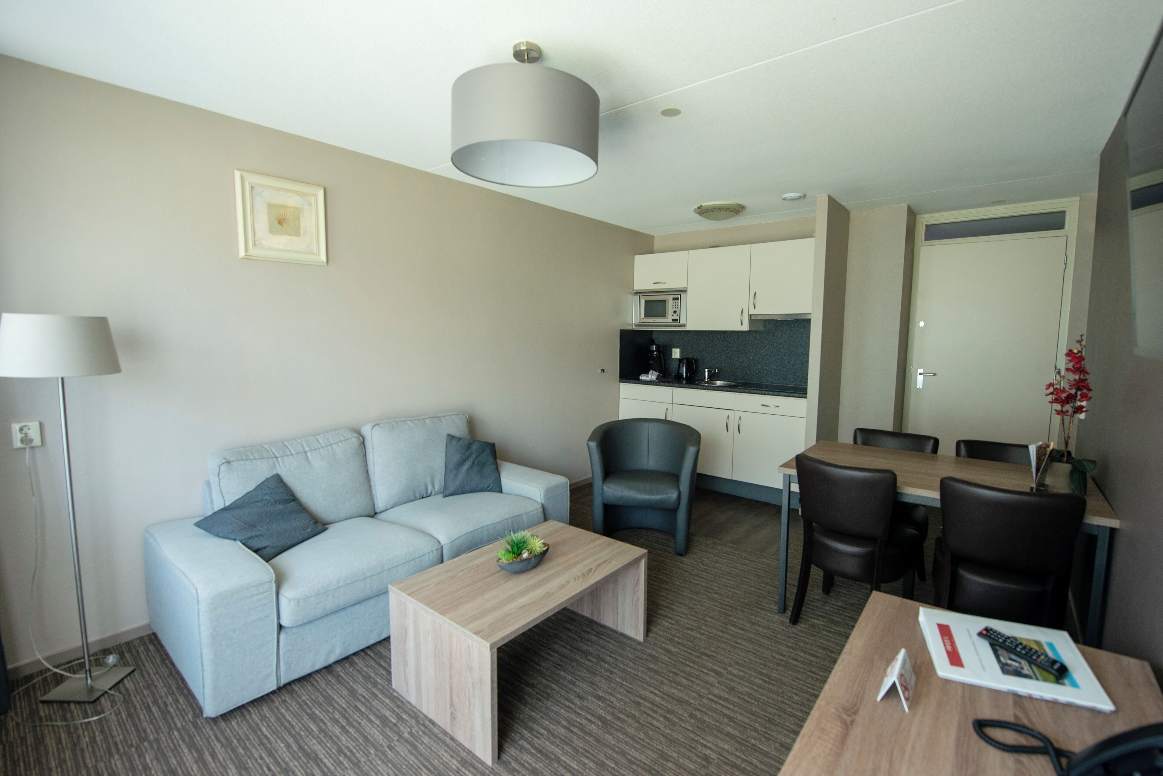 Een moderne hotelkamer met een lichtblauwe bank, een houten salontafel, een eettafel met stoelen, een keukenhoek met kastjes en een magnetron, en een vloerlamp.