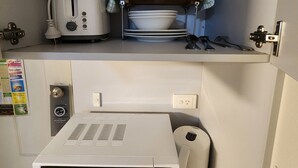 A compact kitchen with a microwave, toaster, and a shelf with cups and plates. - Hume Inn Motel Albury CBD (South Albury)