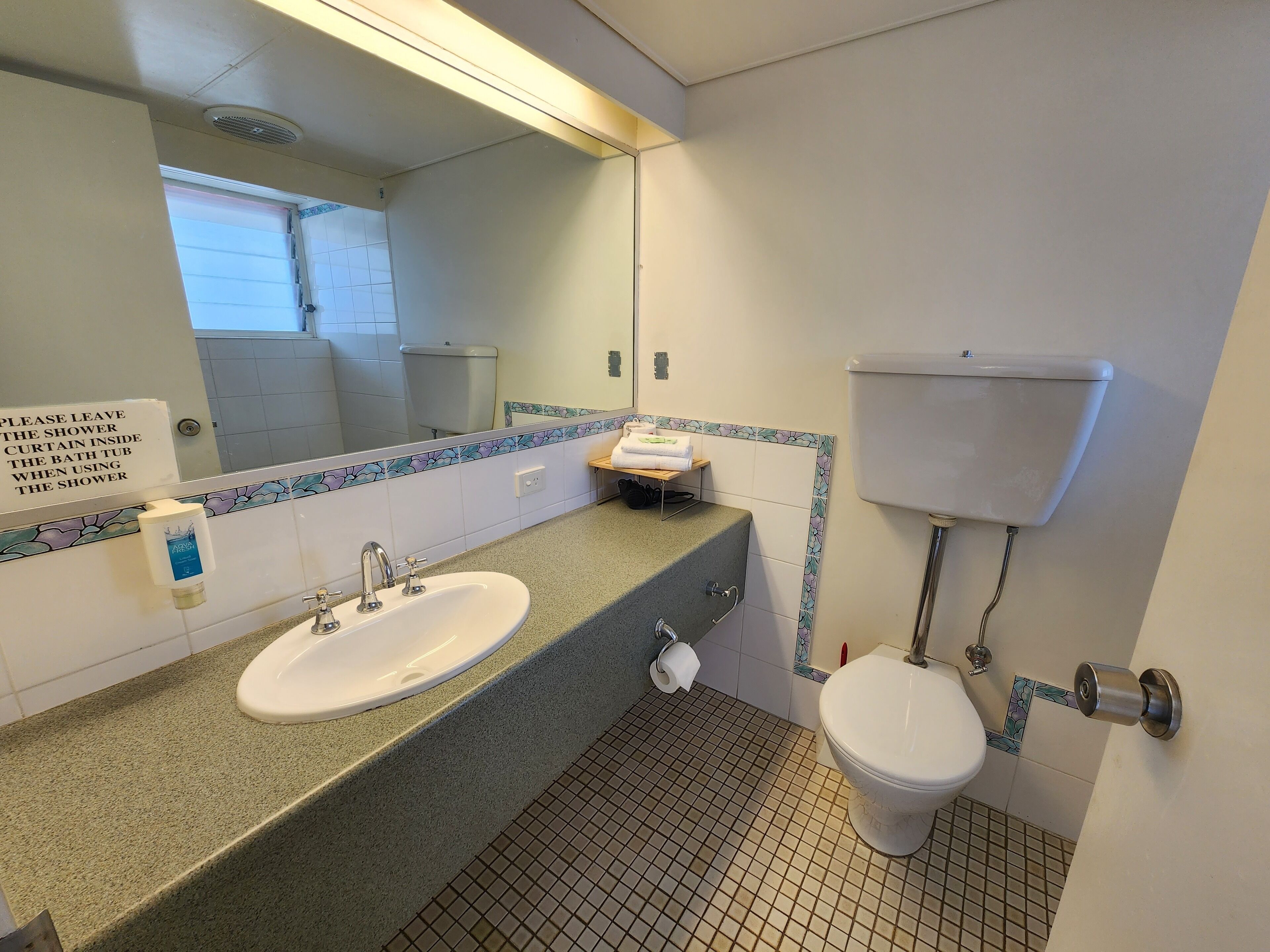 A bathroom with a sink, toilet, and a mirror. A sign instructs to leave the shower curtain inside the shower.