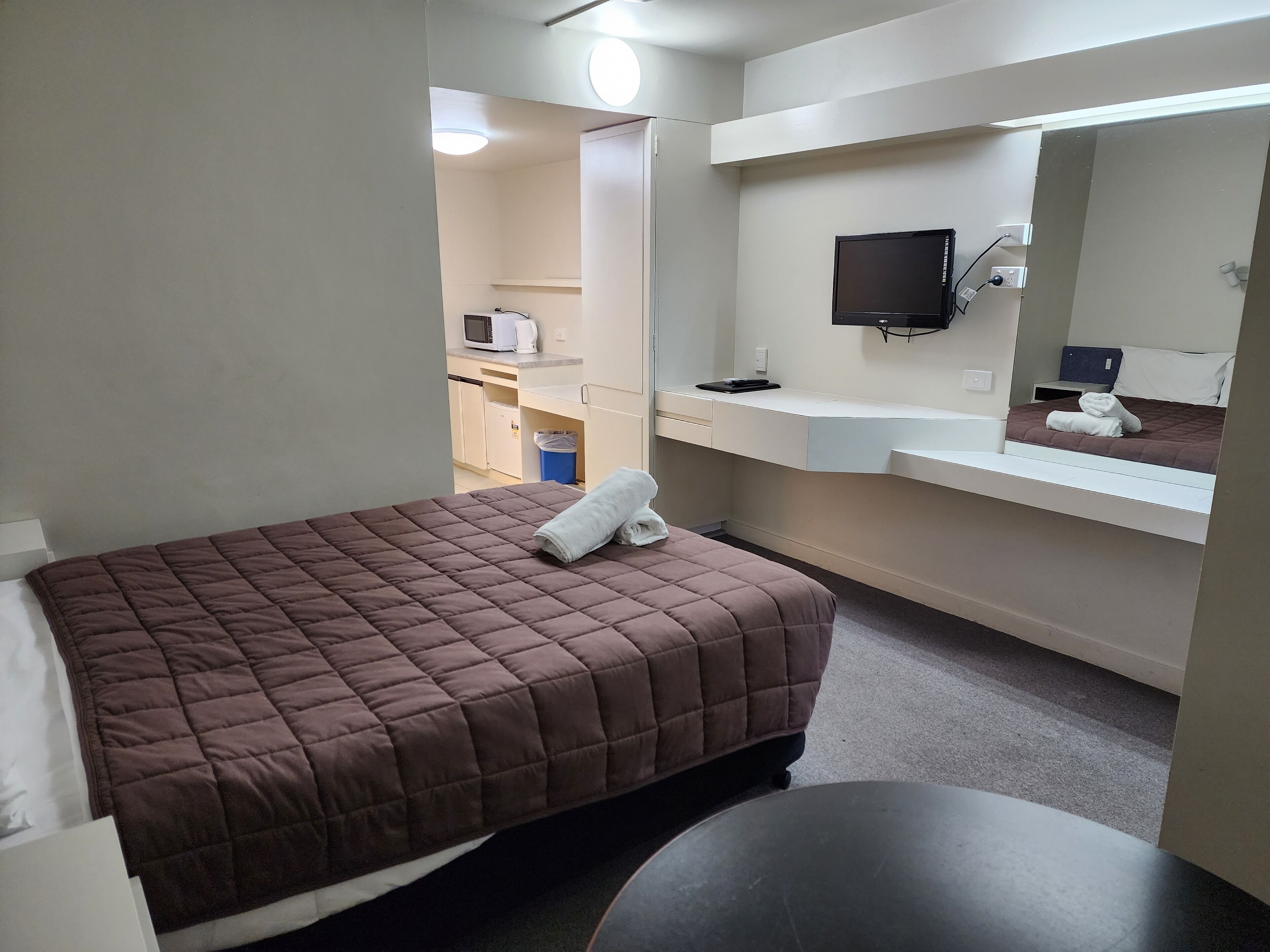 A hotel room with a bed, a small table, a mirror, a TV, and a kitchenette area.