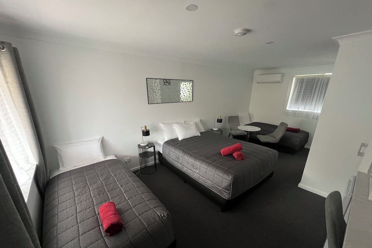 A hotel room with two beds, a small table, a chair, a sofa, and a window with curtains.