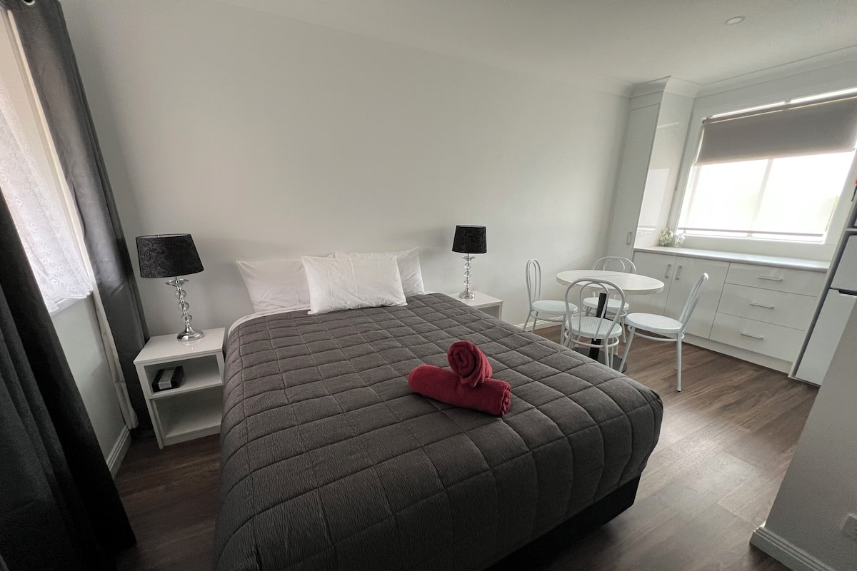 A hotel room with a large bed, two bedside tables with lamps, a small round table with chairs, a kitchenette, and a window with blinds.