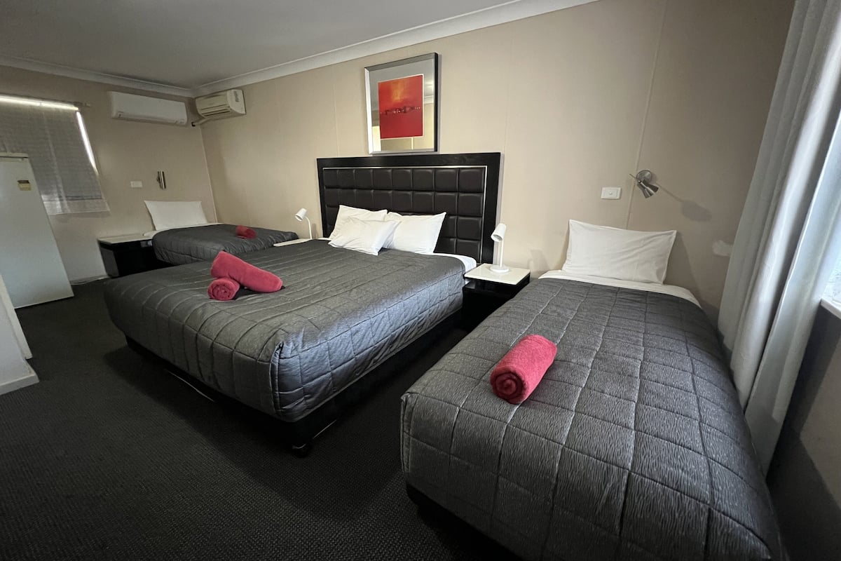 A hotel room with two beds, each with a grey quilted mattress and a black headboard. There is a white bedside table with a lamp, a small nightstand, and a wall-mounted air conditioning unit.