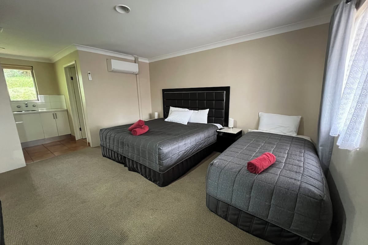 A hotel room with two beds, a small bedside table, a nightstand, a window with curtains, and a kitchenette in the background.