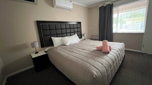 A hotel room with a large bed, a headboard, an air conditioning unit, a nightstand, a lamp, a window with curtains, and a view of greenery outside. - Almond Inn Motel (East Tamworth)