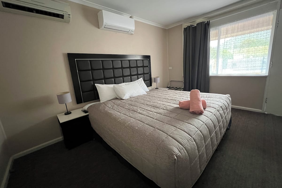A hotel room with a large bed, a headboard, an air conditioning unit, a nightstand, a lamp, a window with curtains, and a view of greenery outside.