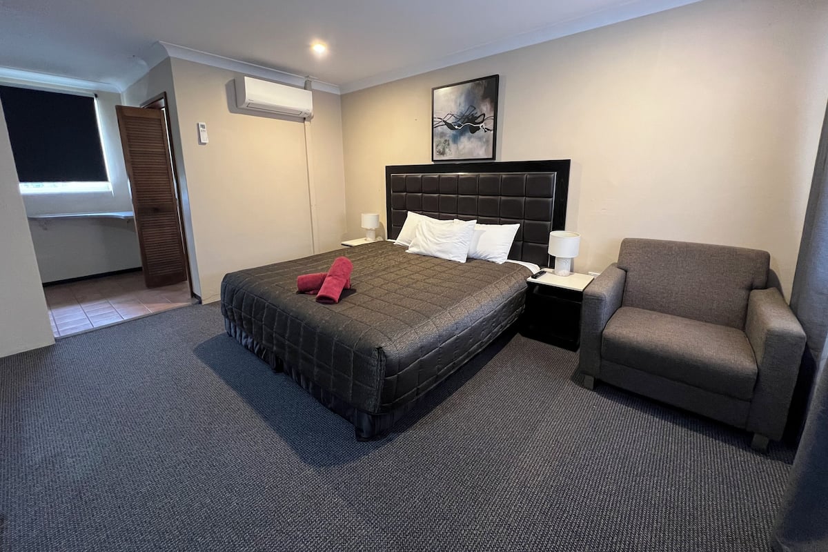 A hotel room with a large bed, a chair, a nightstand, a lamp, and a painting on the wall.