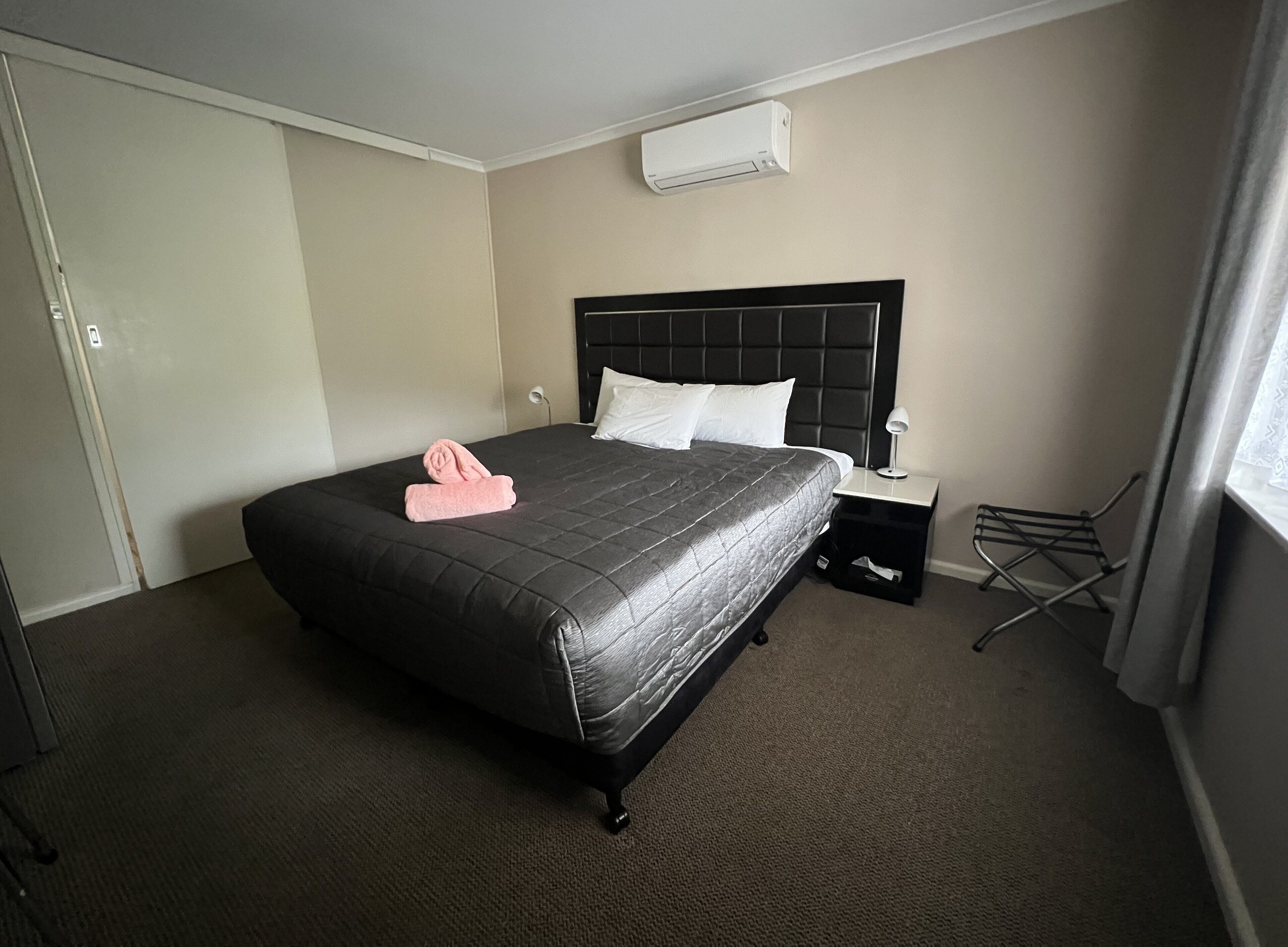 Habitación de hotel con una cama grande, una mesita de noche con lámpara, una silla pequeña y una ventana con cortinas.