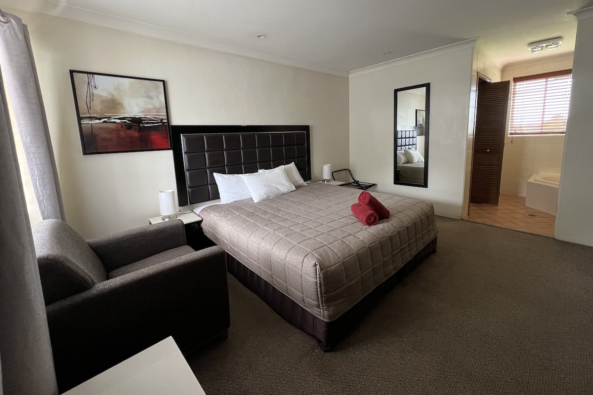 A hotel room with a bed, a chair, a bedside table, a mirror, and a bathroom door.