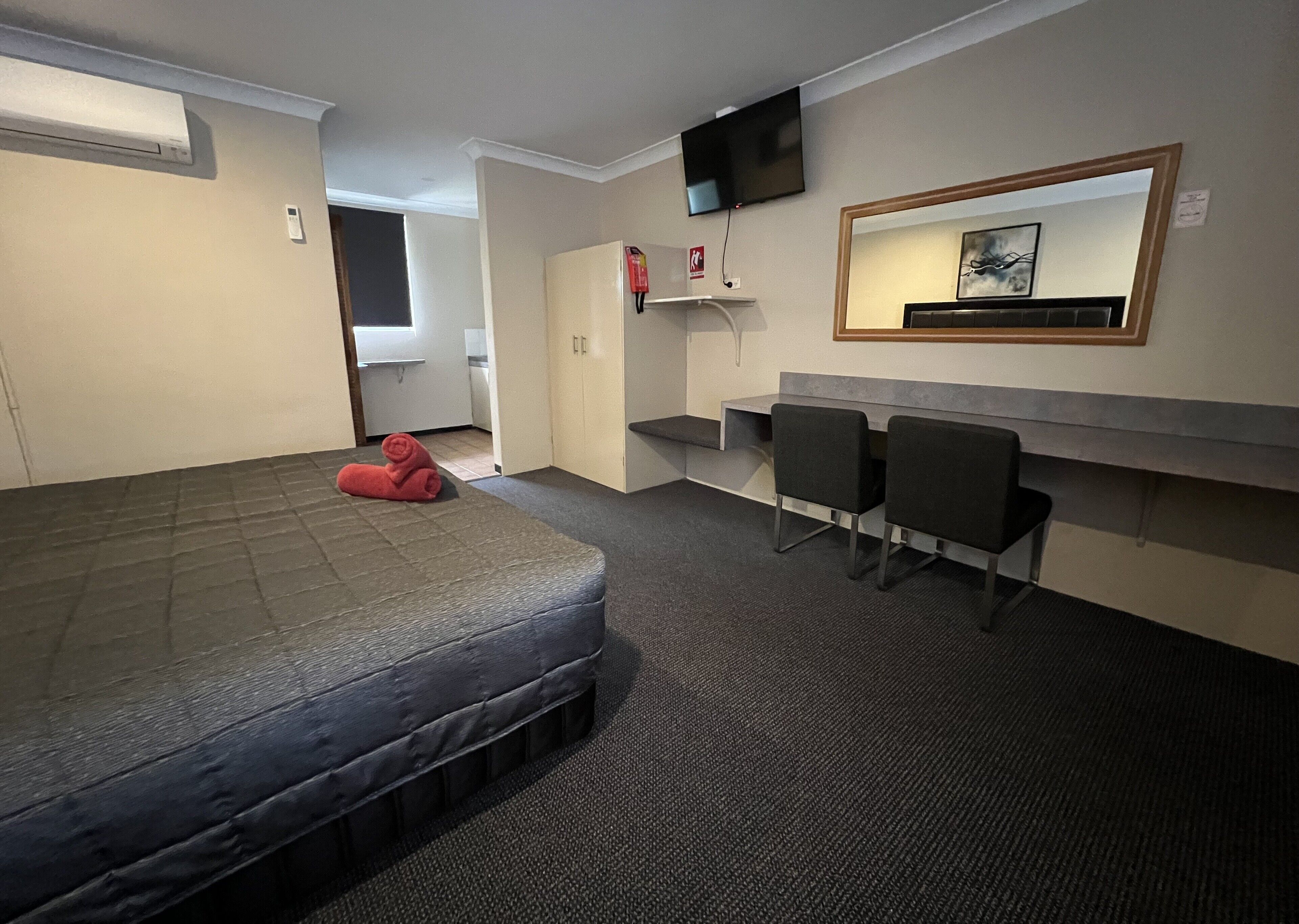 A room with a bed, two chairs, a desk, a TV, and a small shelf with a picture.