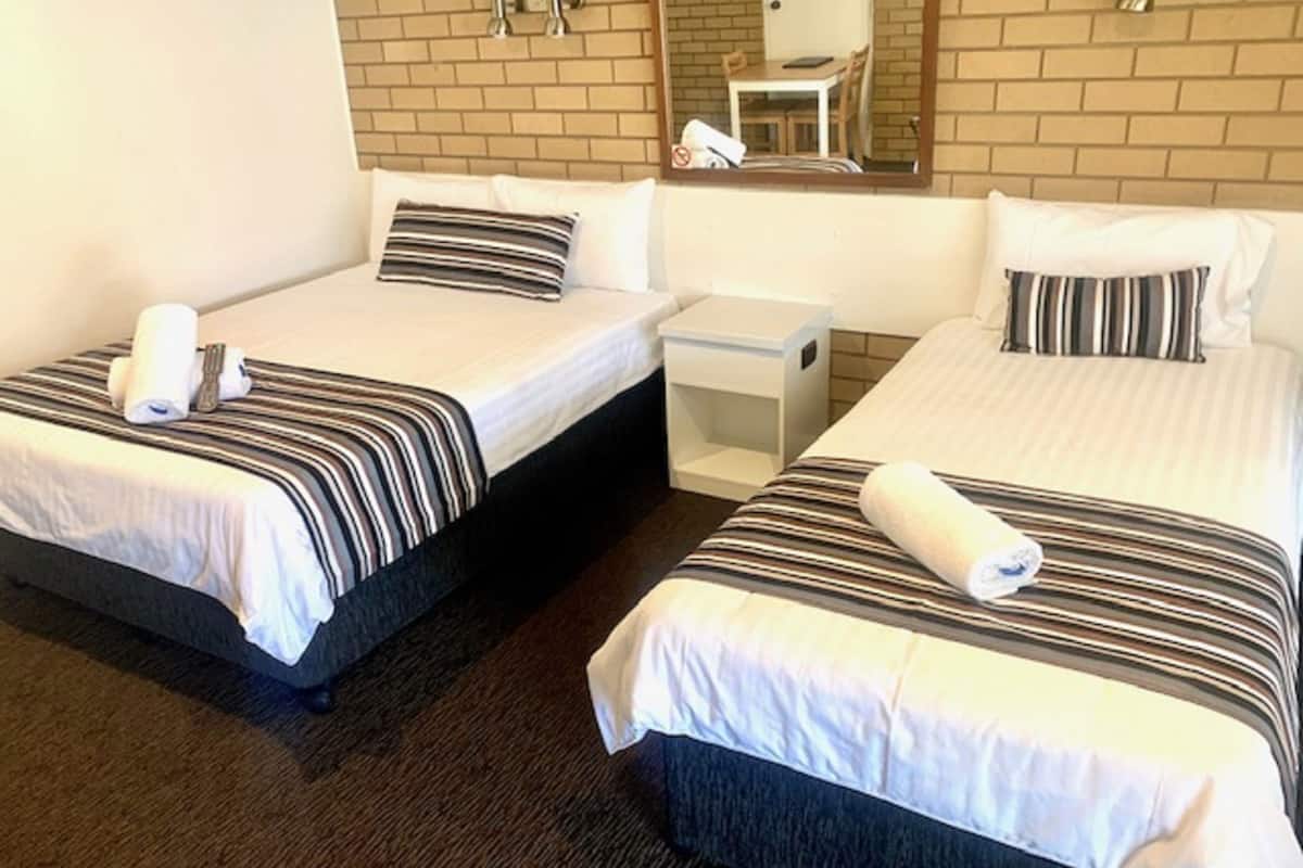 A hotel room with two single beds, each with striped bedding and a pillow. There is a small bedside table with a lamp and a chair visible in the reflection of a mirror.