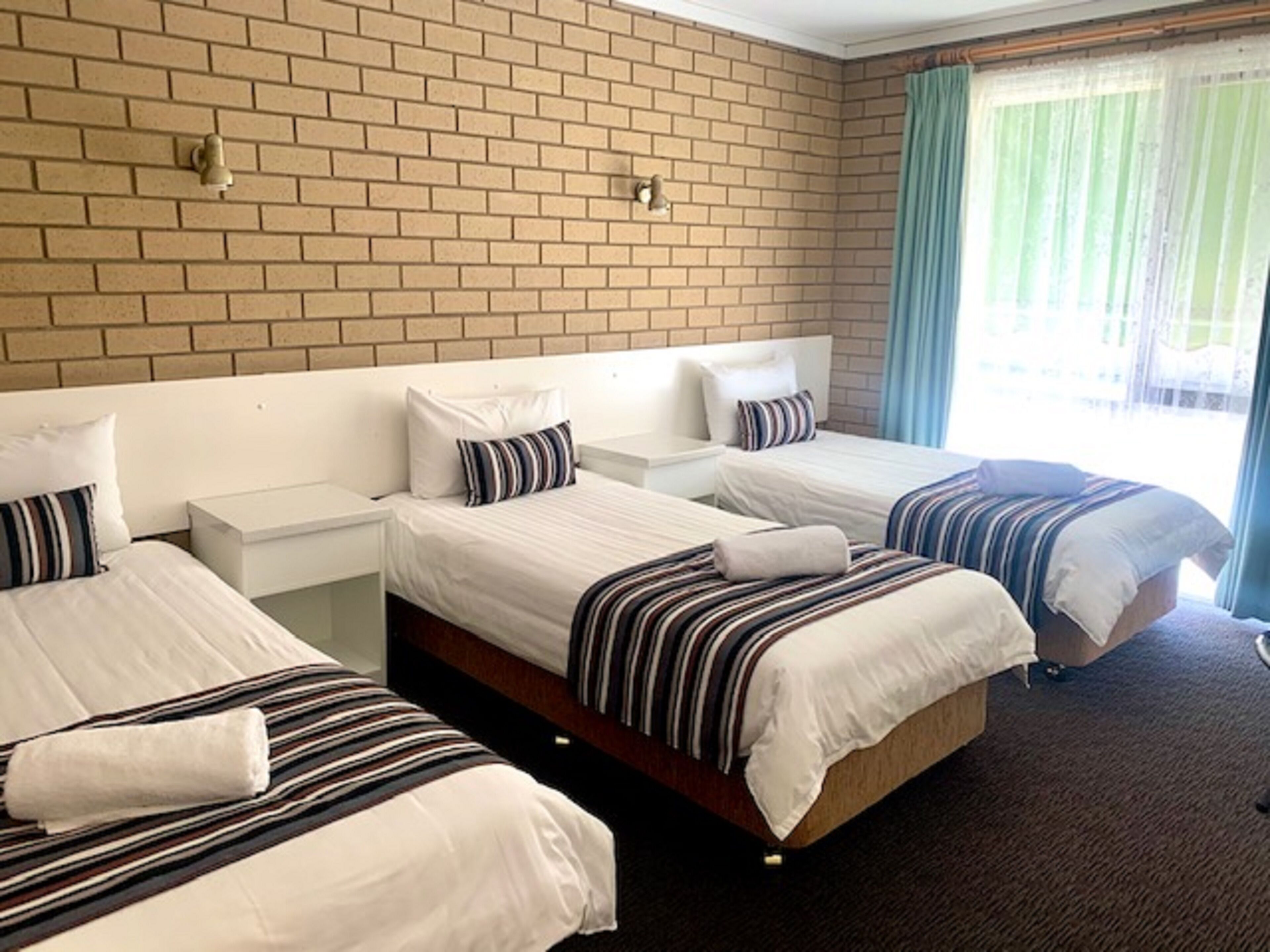Habitación de hotel con dos camas individuales, una pared de ladrillo, una ventana con cortinas y una mesita de noche con lámpara.