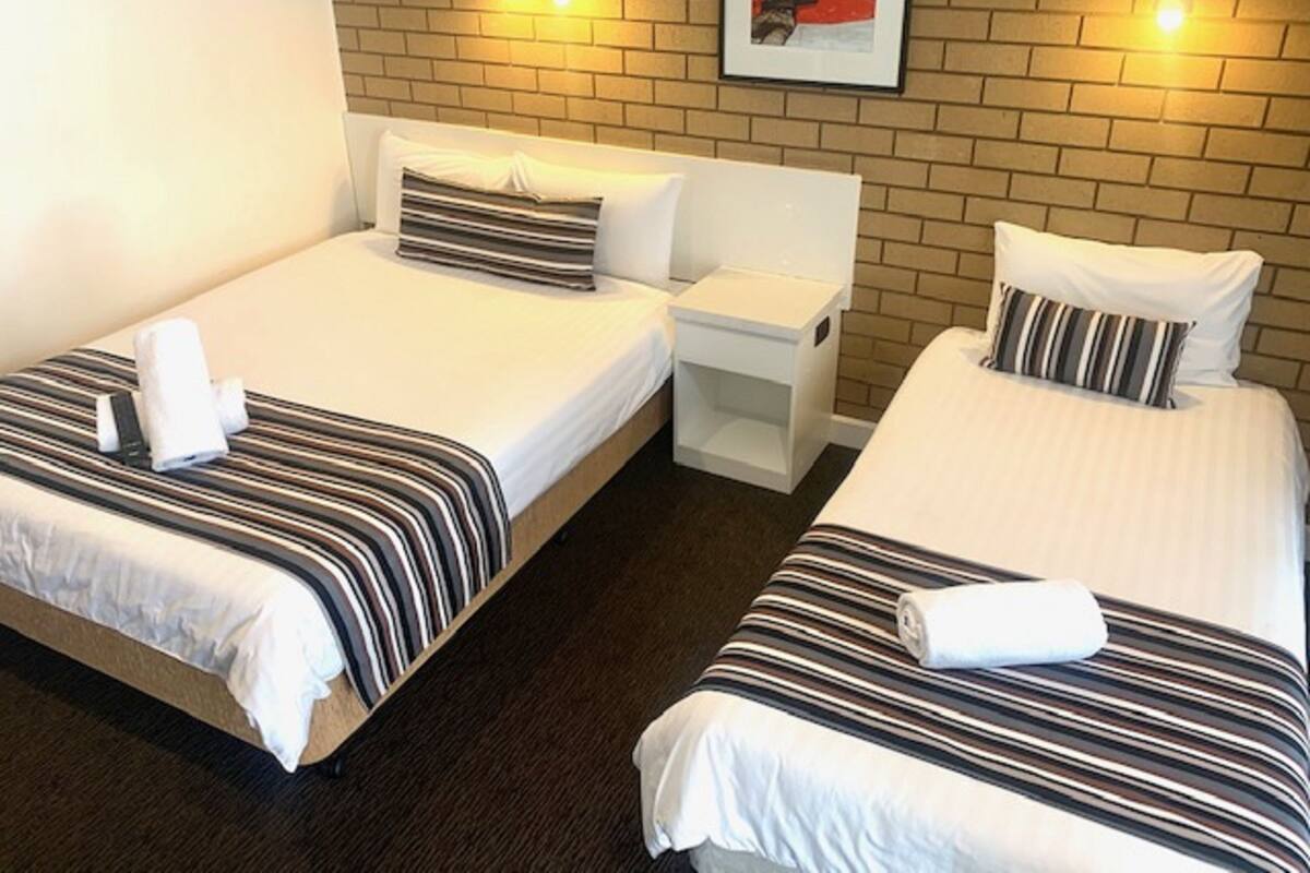 A hotel room with two single beds, each with striped bedding and a white pillow. There is a small white nightstand between the beds, and a framed picture on the wall above the beds.