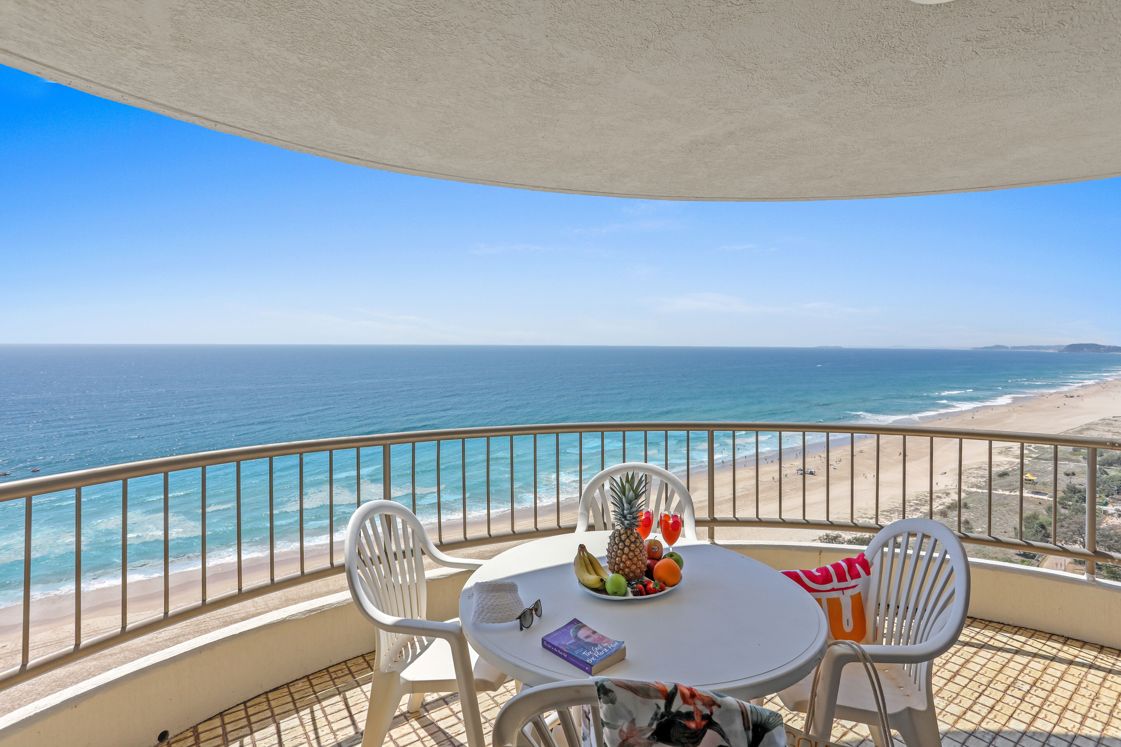 A balcony with a table set with fruit, a book, and a handbag. The view includes a beach and the ocean.