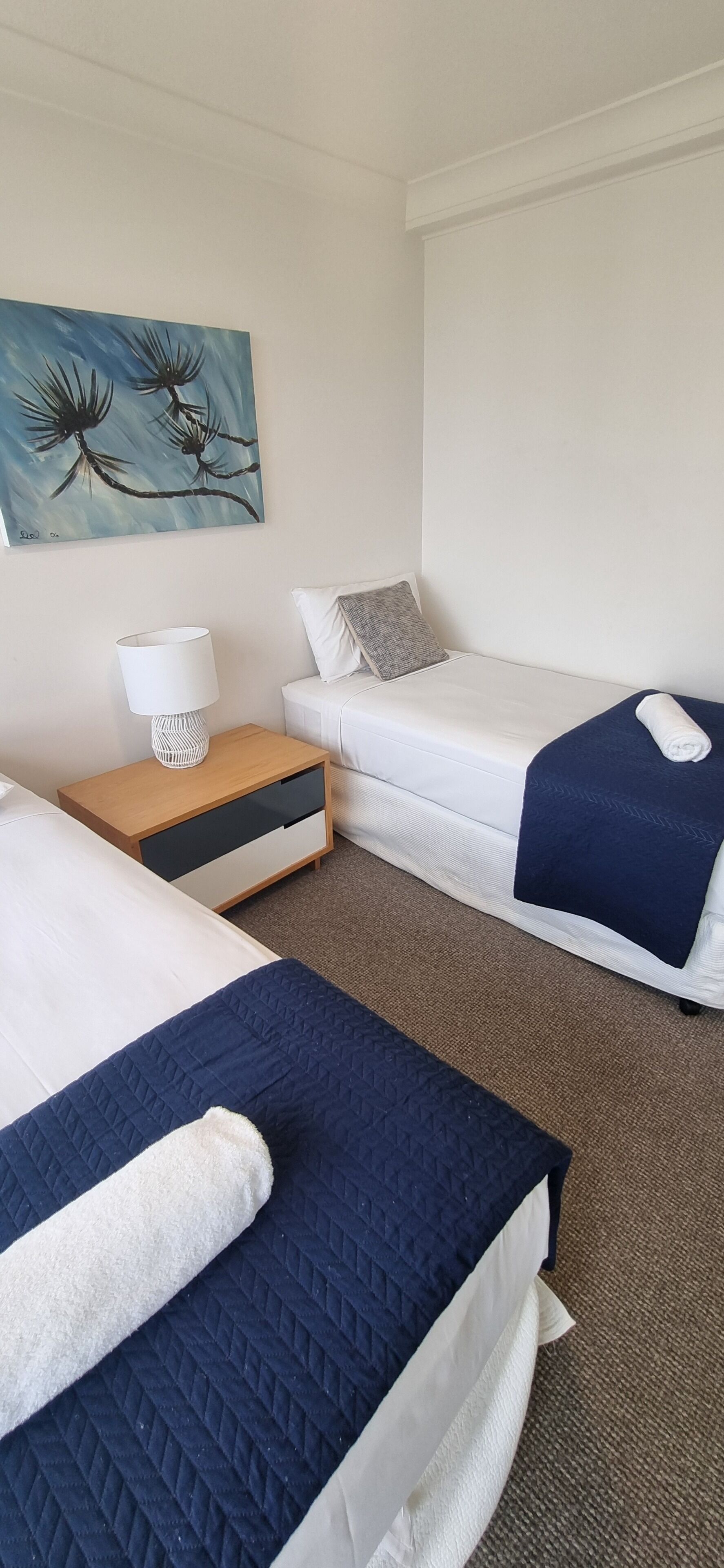 Habitación de hotel con dos camas, una mesita de noche con lámpara, un cuadro en la pared y suelo alfombrado.