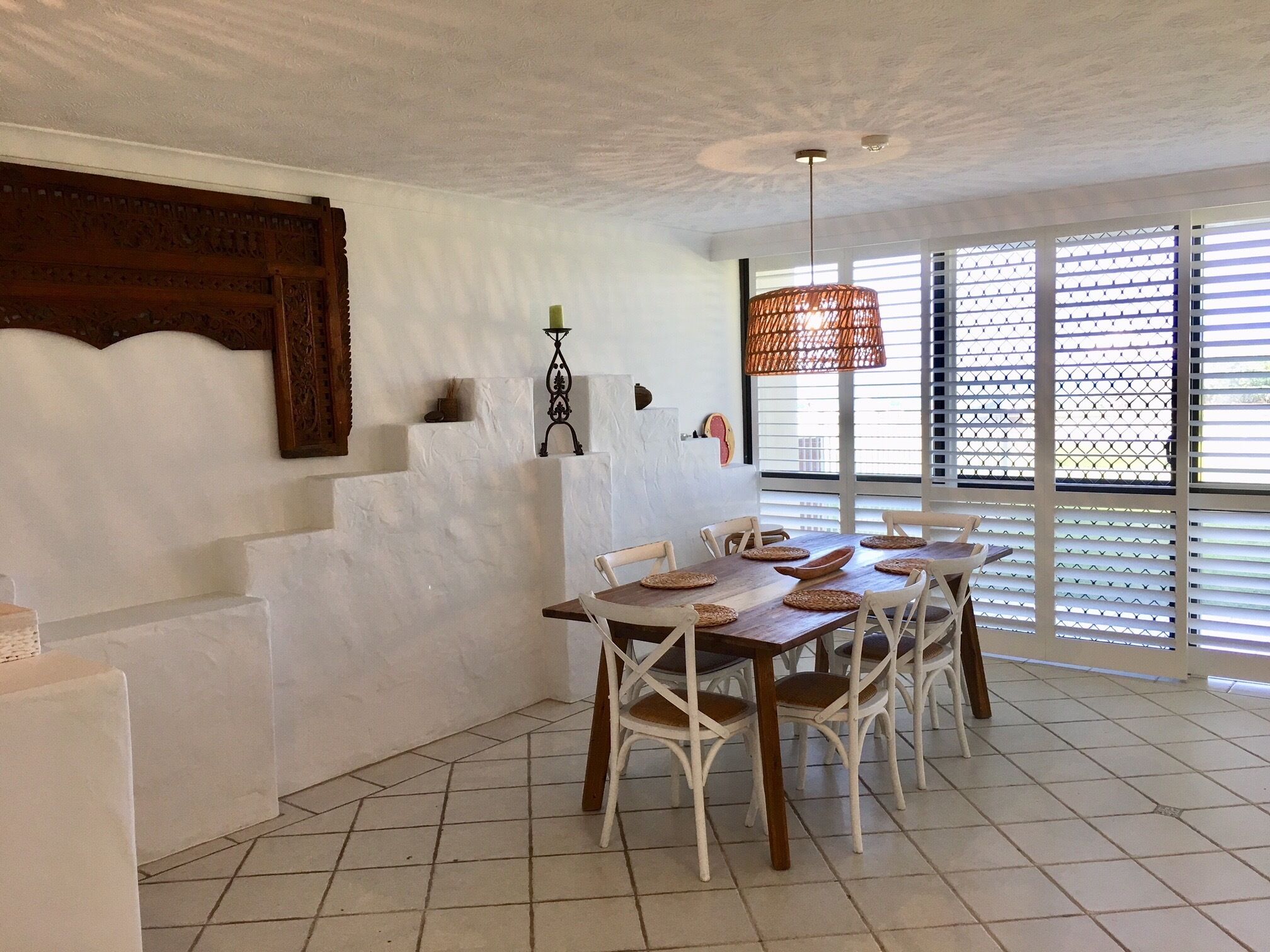 A dining area with a white tiled floor, white chairs, a wooden table, and a hanging rattan light fixture.