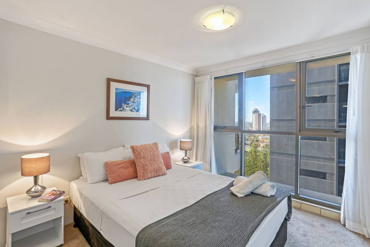 Een moderne hotelkamer met een groot bed, een nachtkastje met een lamp, een ingelijst schilderij aan de muur, en een uitzicht op de stad door een groot raam.