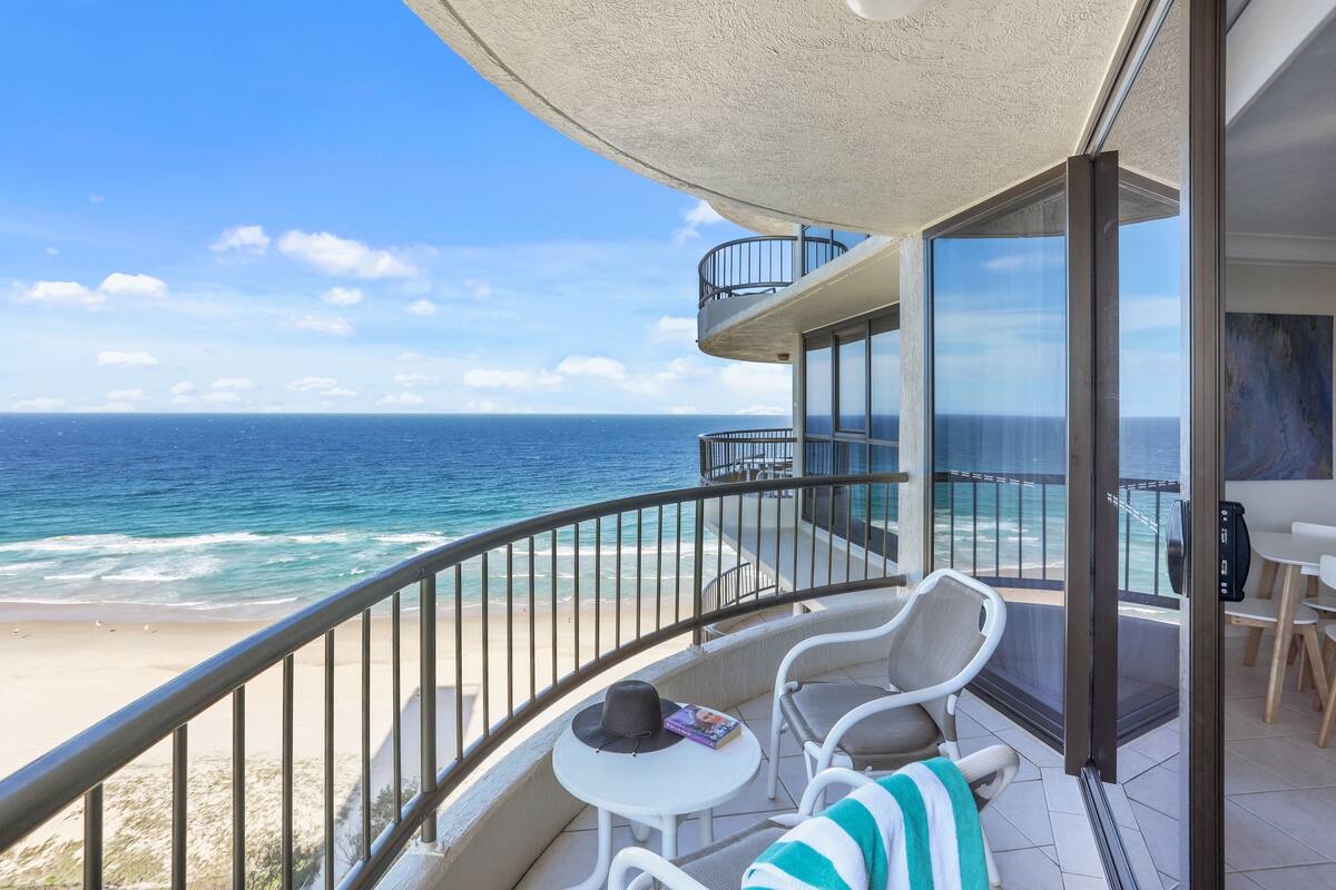 Een balkon met uitzicht op de oceaan, een tafel met een hoed en een boek, en een stoel met een gestreepte handdoek.