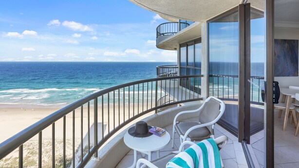 A balcony with a view of the ocean, a table with a hat and a book, and a chair with a striped towel.