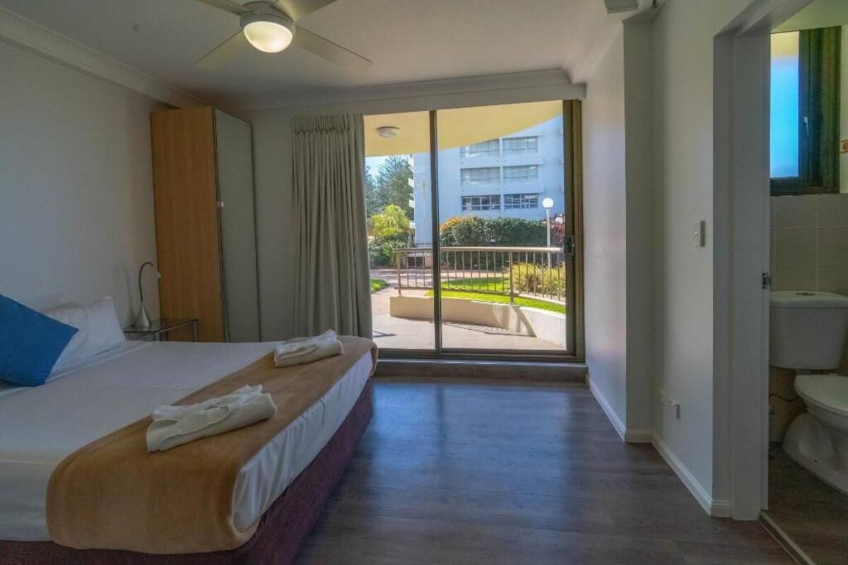 Een hotelkamer met een bed, een badkamer en een balkon met uitzicht op gebouwen en groen.