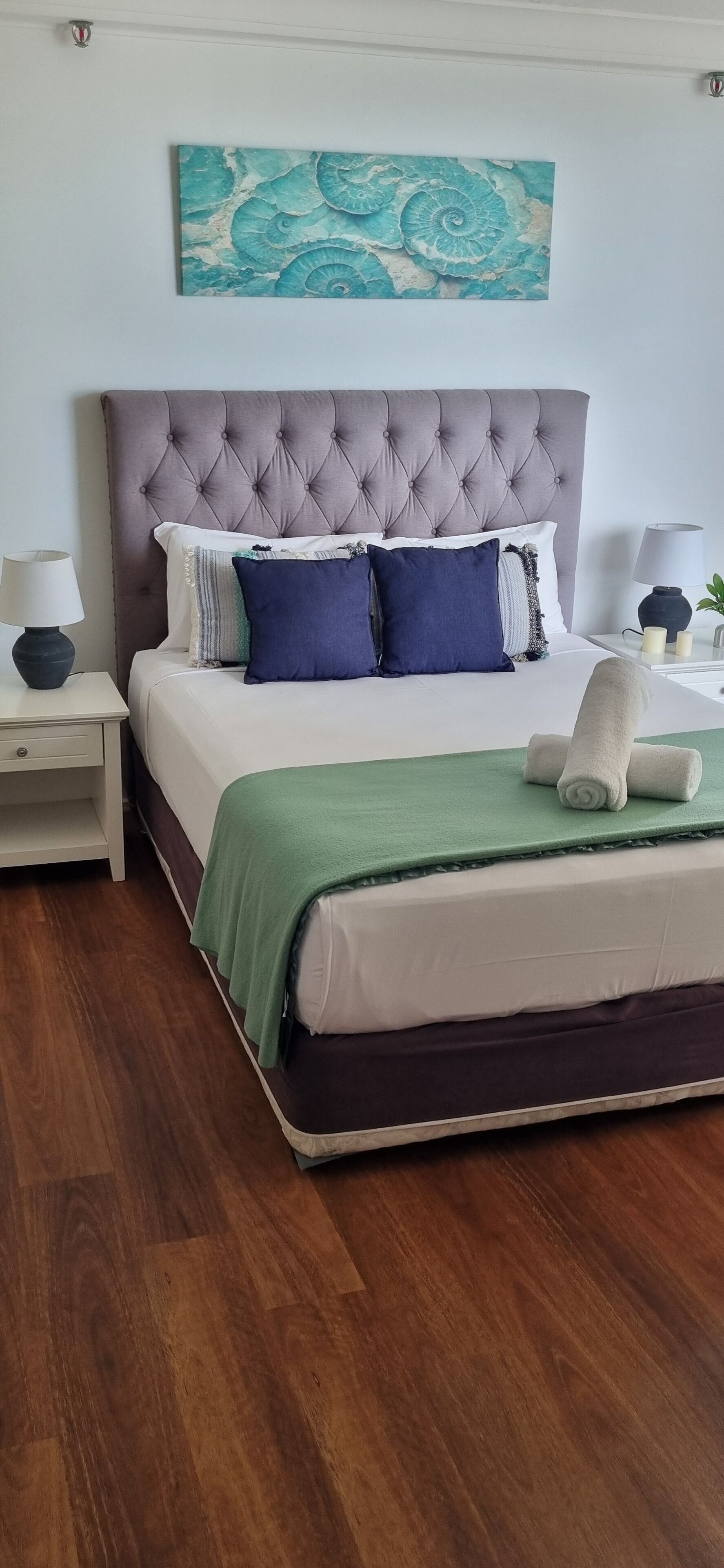 A neatly made bed with a tufted headboard, a green throw blanket, and rolled towels.