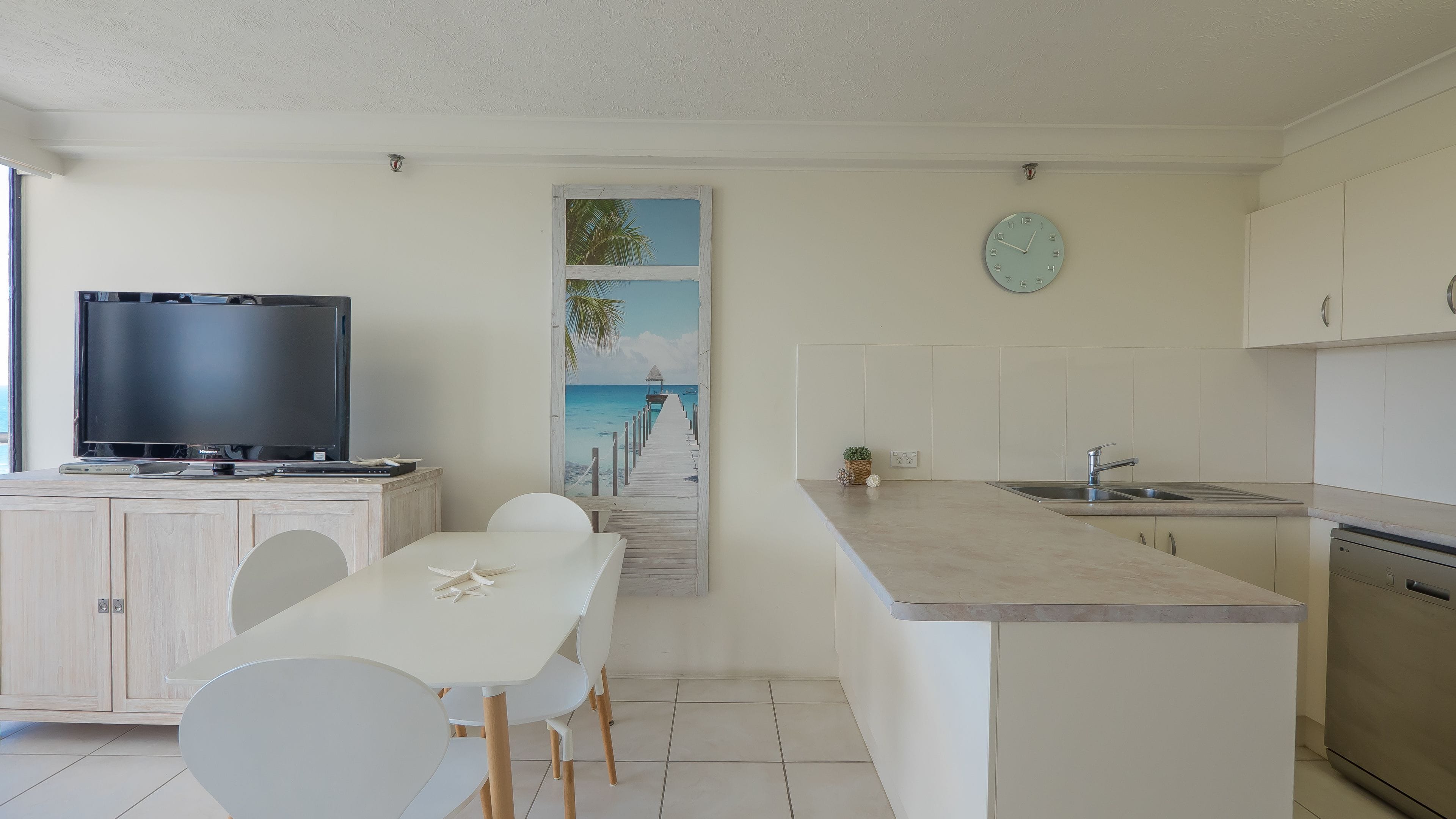 Una cocina moderna con una isla central, un comedor con sillas blancas, un televisor de pantalla plana y un reloj de pared.