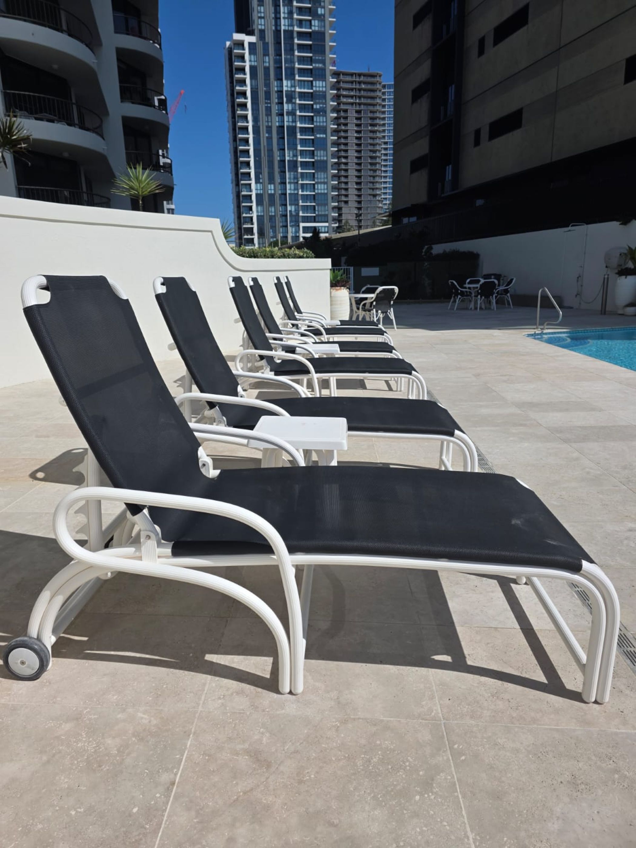 A row of black and white lounge chairs by a swimming pool with a cityscape in the background.