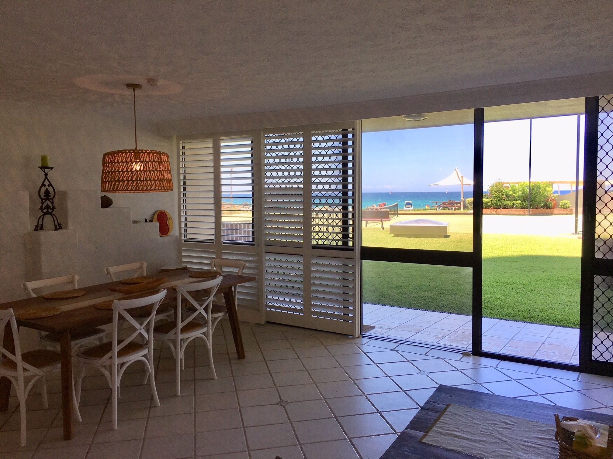 Un comedor con una mesa de madera y sillas blancas, una lámpara colgante y vistas a un área exterior con césped, una playa y una tienda de campaña.