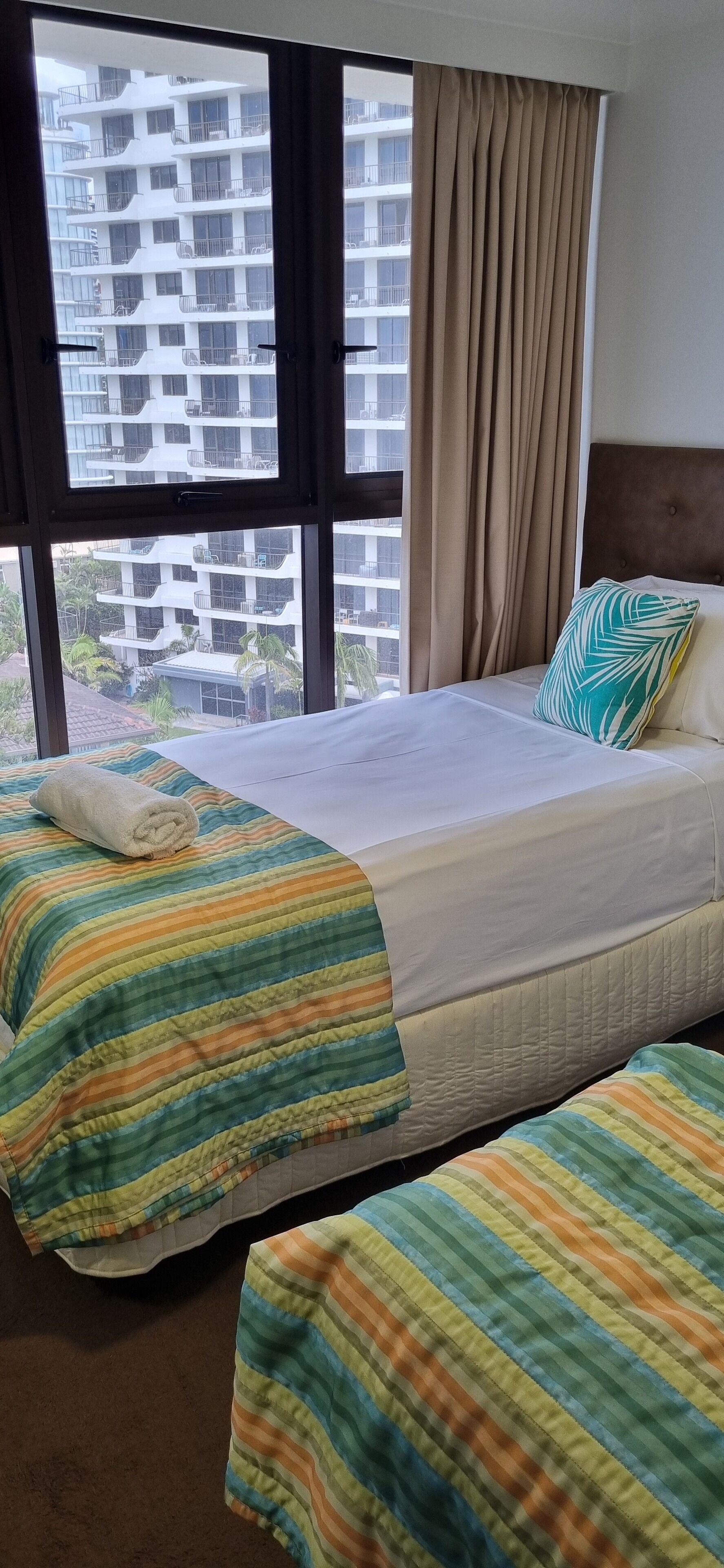 Une chambre d’hôtel avec un grand lit, deux oreillers, une couverture à rayures et une vue sur un immeuble par une fenêtre ornée de rideaux.