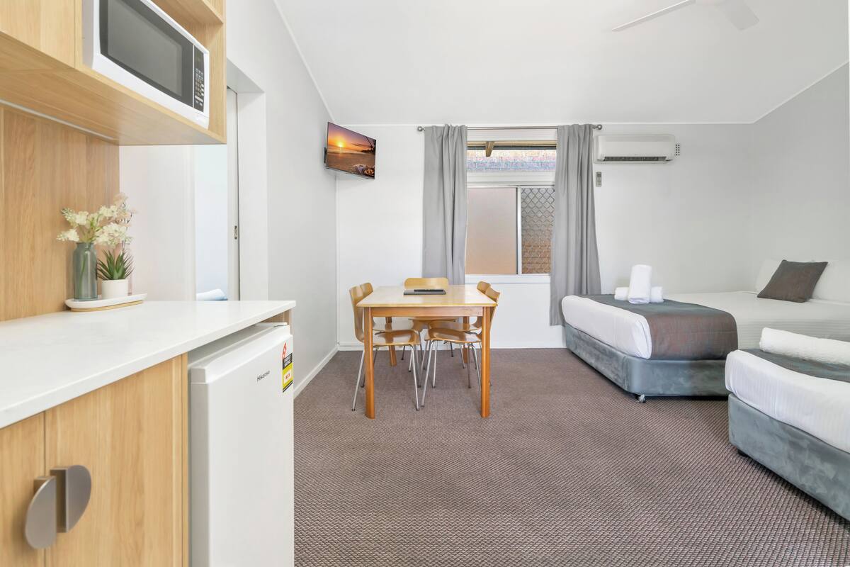 A compact hotel room with a bed, a desk with a chair, a microwave, a TV, and a ceiling fan.