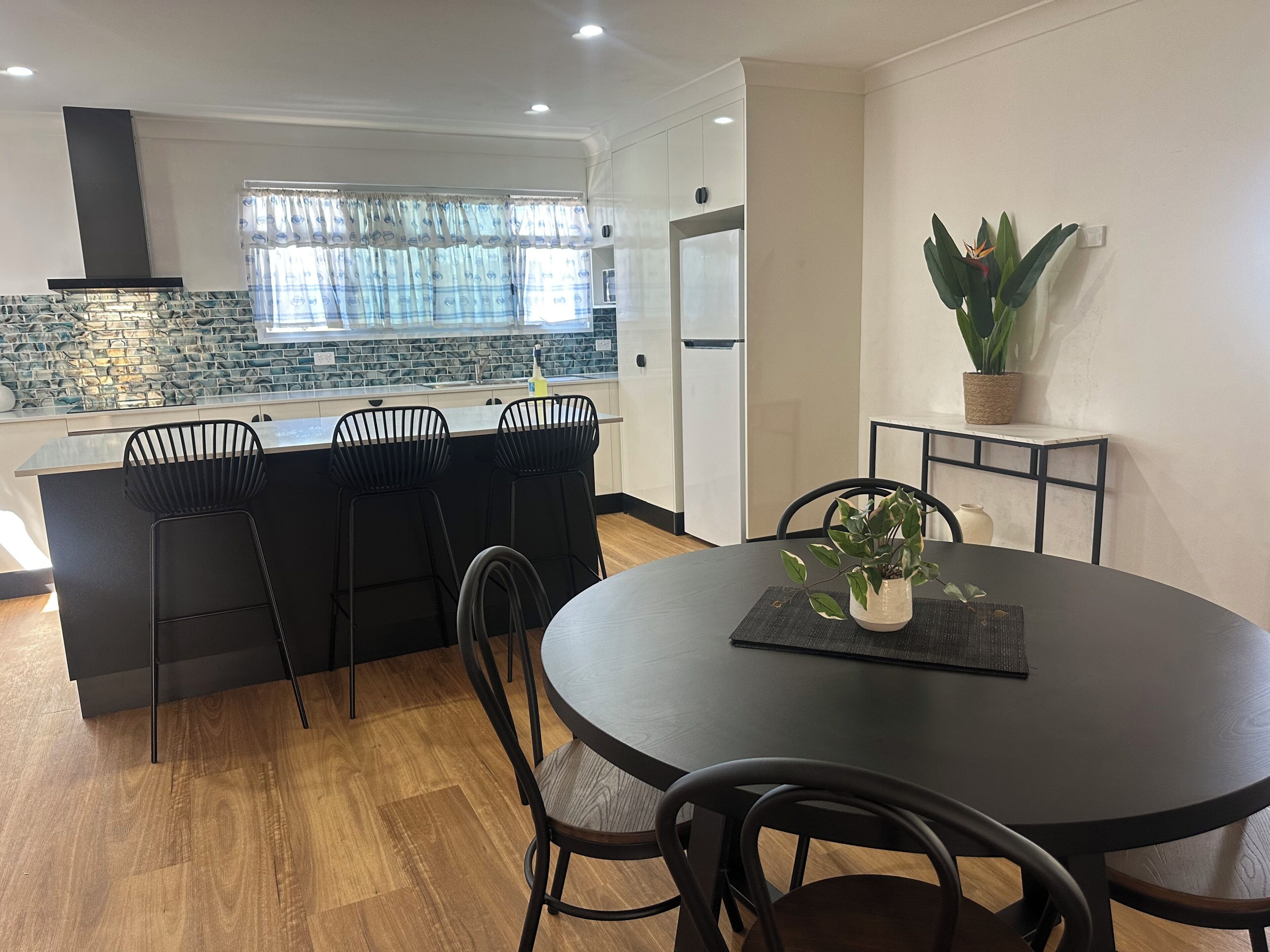 A modern kitchen with a stone backsplash, a central island with bar stools, a dining table with chairs, and a potted plant on a console table.
