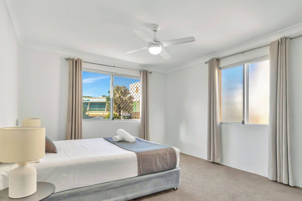 A hotel room with a bed, a ceiling fan, a window with curtains, and a view of buildings outside.