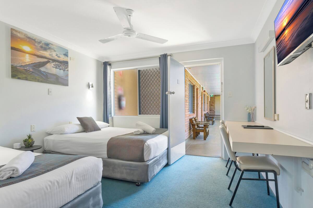 A hotel room with two beds, a desk, and a chair. There is a ceiling fan and a painting on the wall.