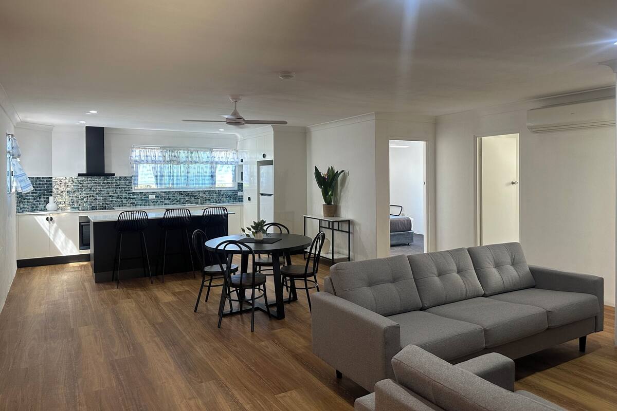 A modern living room with a grey sofa, a dining area with a table and chairs, a kitchen with a built-in island, and a ceiling fan.
