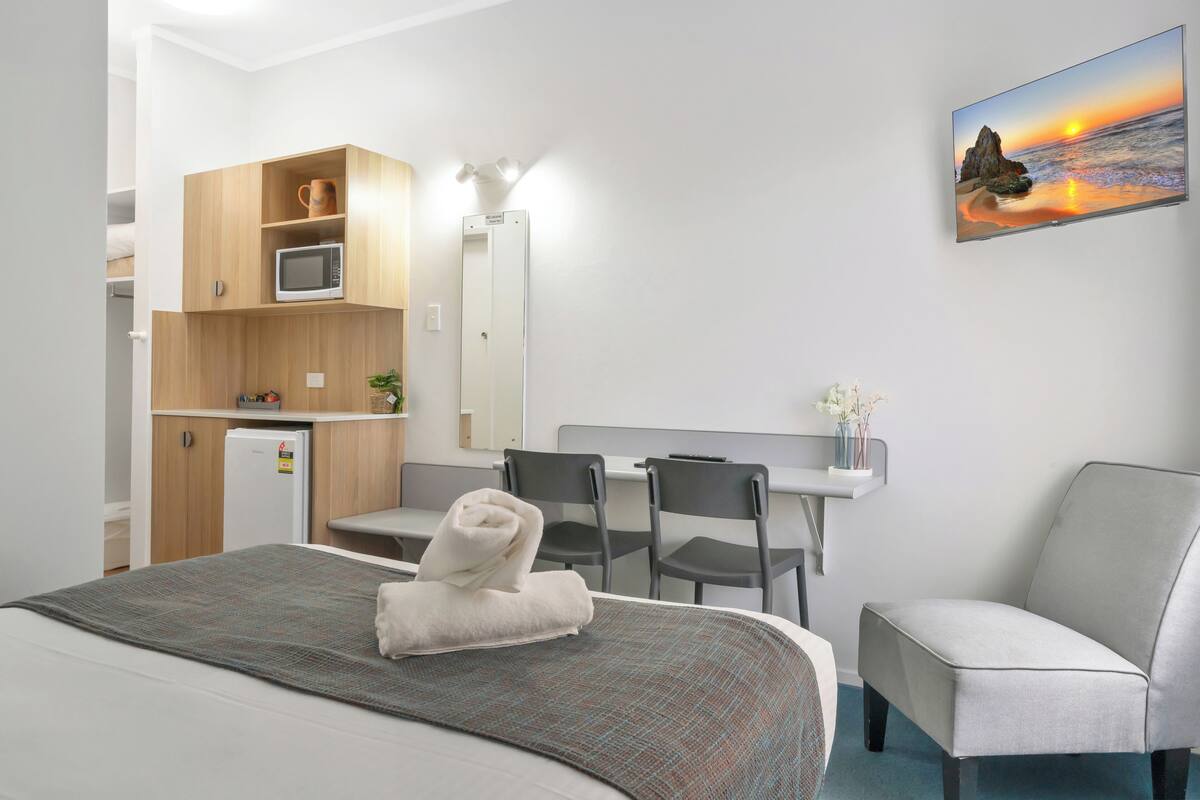 A hotel room with a bed, a desk, a chair, a microwave, a refrigerator, a mirror, and a painting of a sunset.