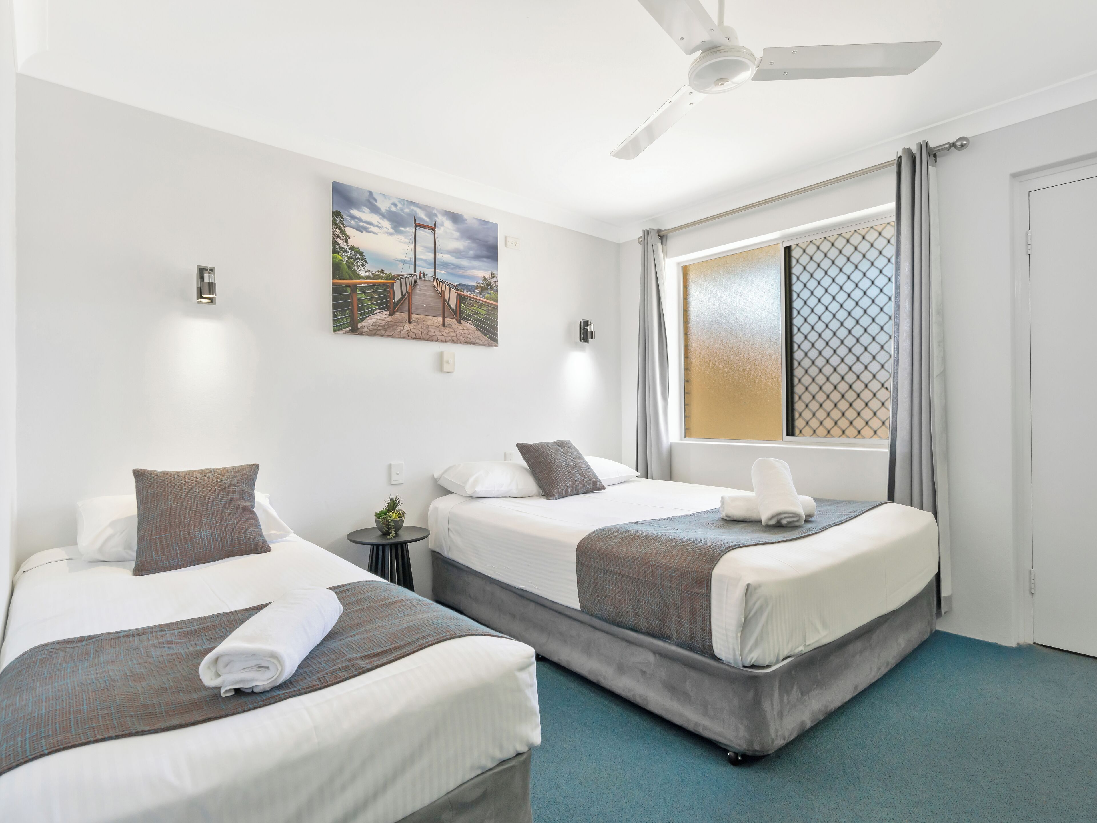 A hotel room with two beds, a ceiling fan, a window with curtains, and a painting on the wall.