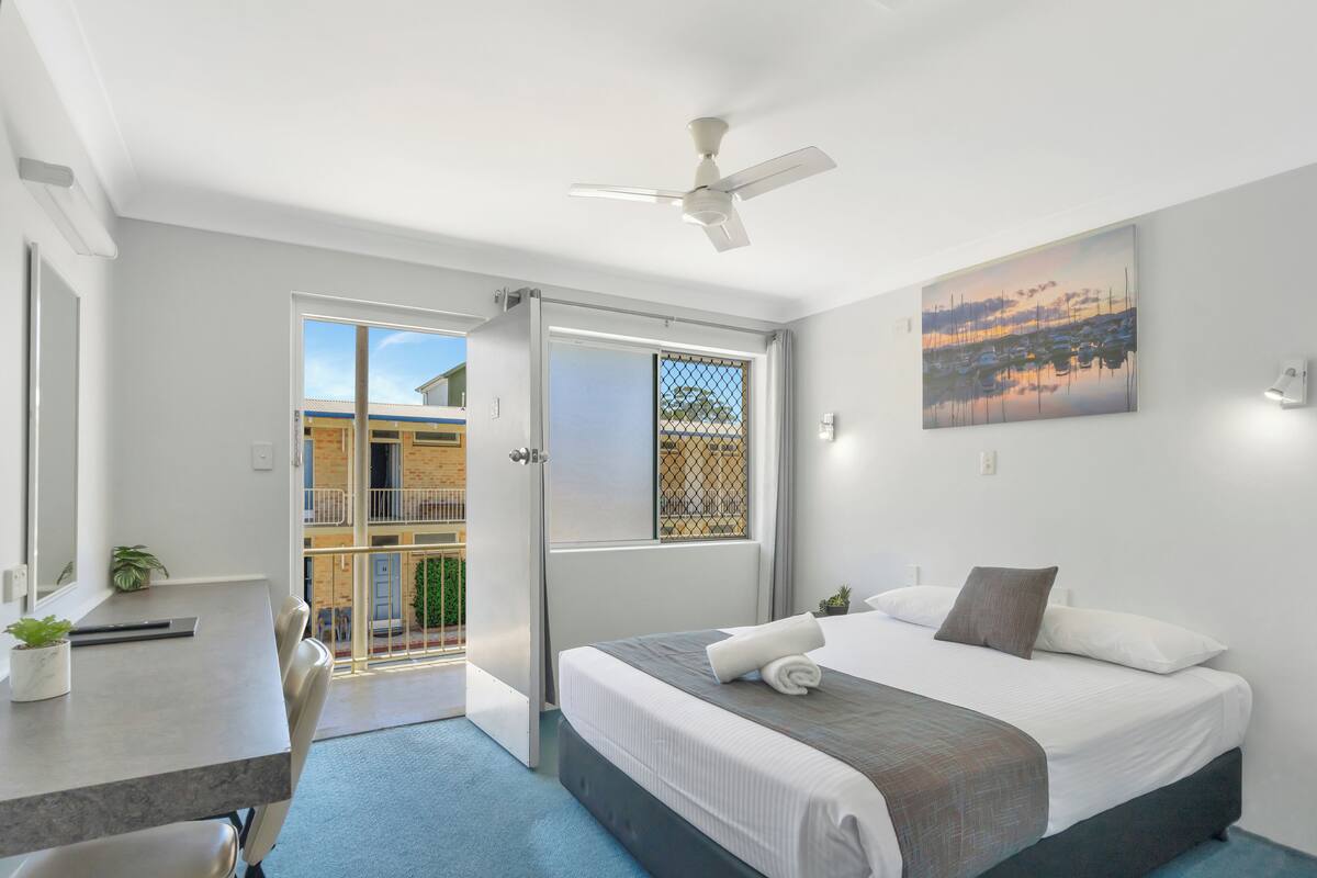 A modern hotel room with a bed, a desk, and a chair. There is a balcony with a view of buildings and a fence.