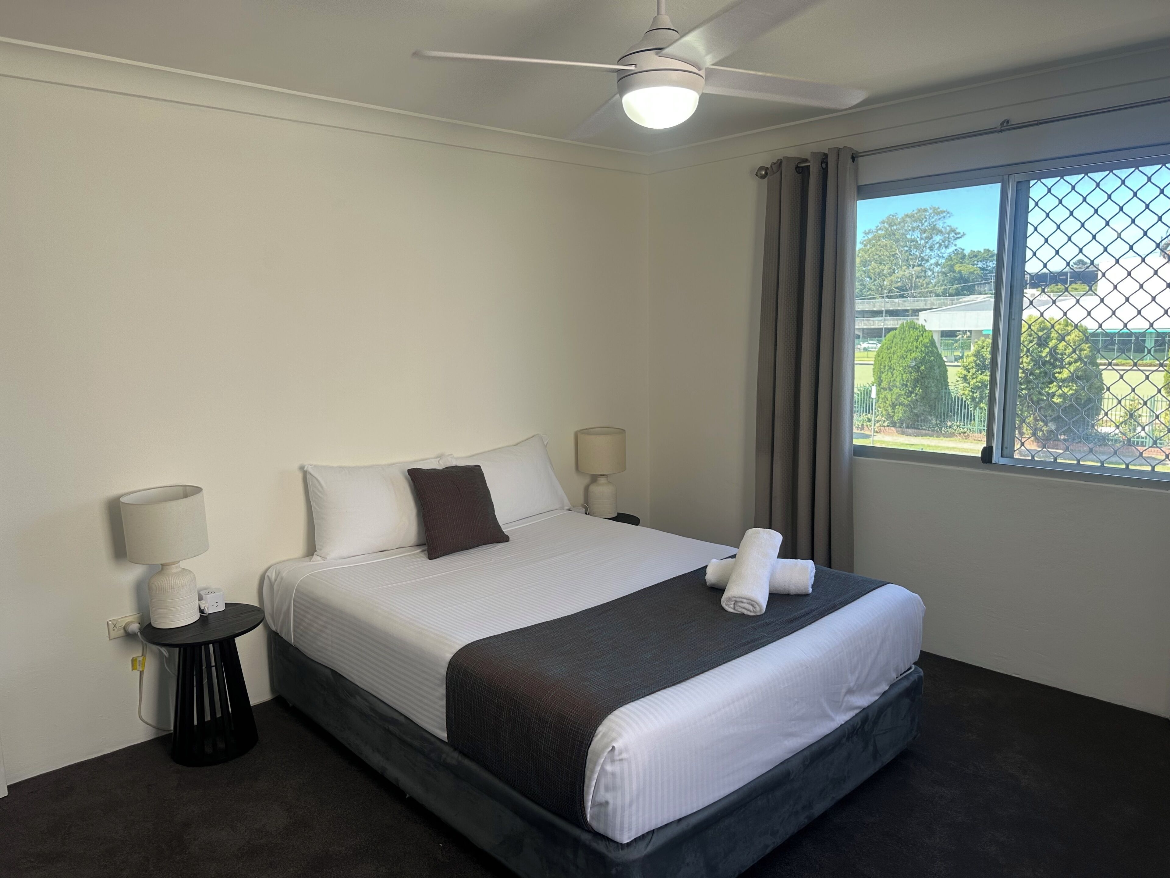 A hotel room with a bed, two bedside tables, a ceiling fan, a window with a view, and a power outlet.
