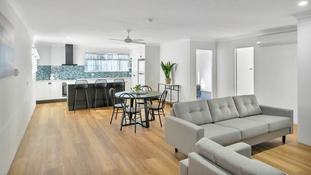 Una sala de estar moderna con un sofá gris, una zona de comedor con mesa y sillas, una cocina con salpicadero azul y un ventilador de techo.