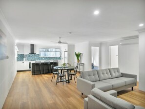 Una sala de estar moderna con un sofá gris, una zona de comedor con mesa y sillas, una cocina con salpicadero azul y un ventilador de techo.