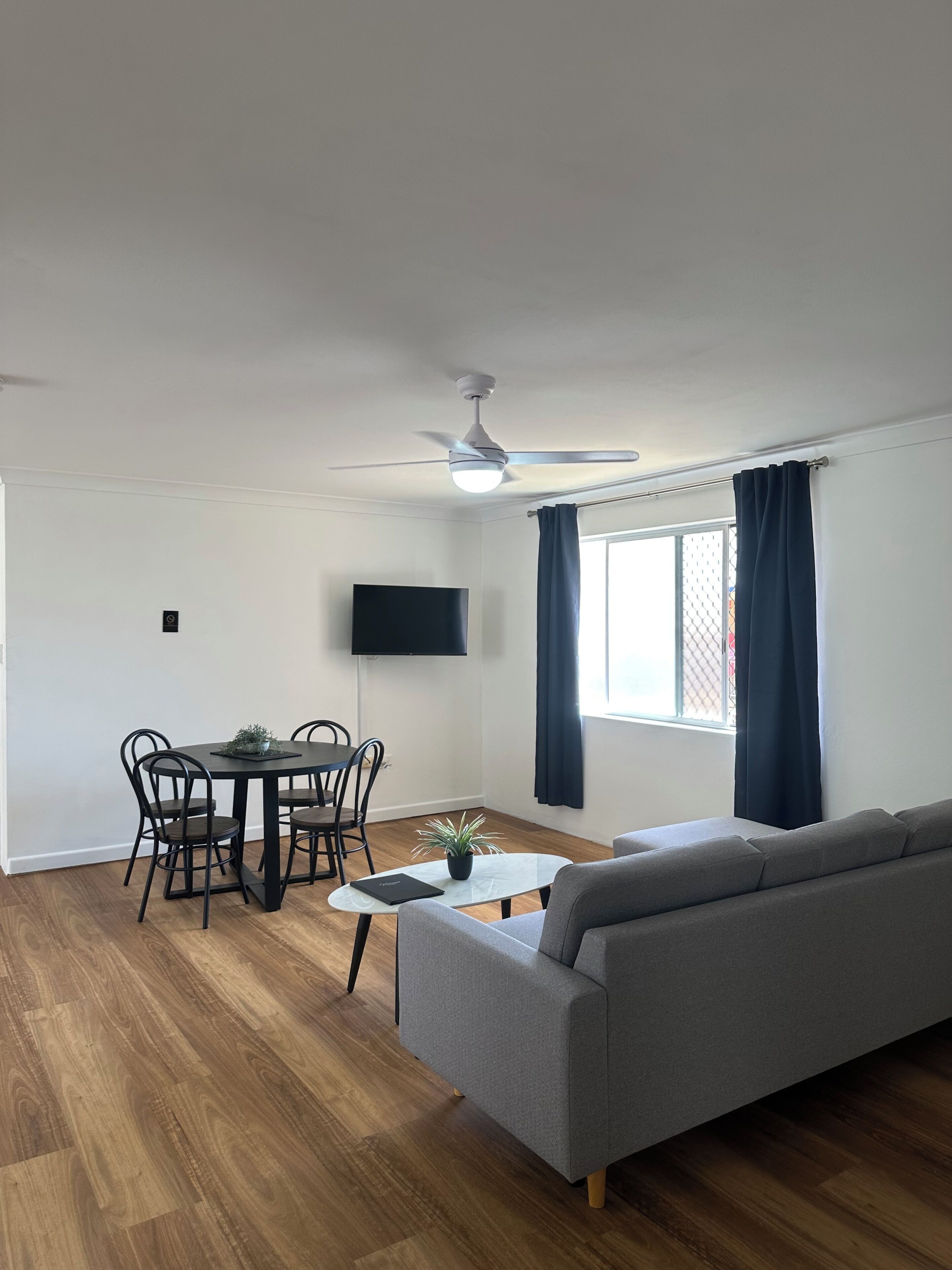 Uma sala de estar moderna com um sofá cinzento, uma mesa de jantar com cadeiras, um ventilador de teto, uma televisão de tela plana e uma janela com cortinas.