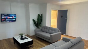 Una sala de estar moderna con un sofá gris, una mesa de centro con una planta, y un televisor montado en la pared mostrando varios canales.