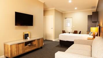 Une chambre d’hôtel équipée d’un téléviseur à écran plat fixé au mur, d’une console en bois, d’un lit recouvert d’une couette blanche, d’un canapé et d’un bureau avec une lampe.
