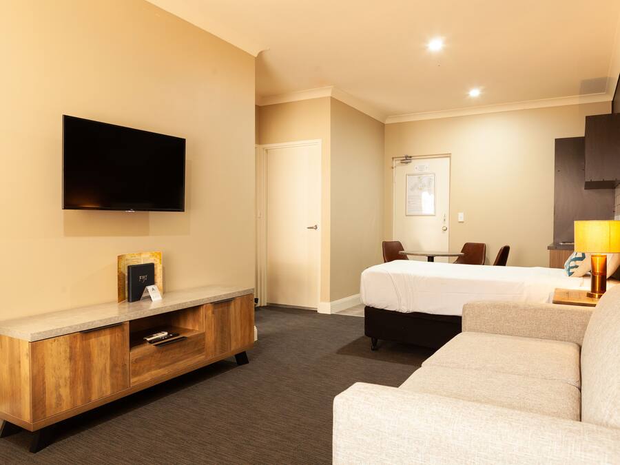 Une chambre d’hôtel équipée d’un téléviseur à écran plat fixé au mur, d’une console en bois, d’un lit recouvert d’une couette blanche, d’un canapé et d’un bureau avec une lampe.