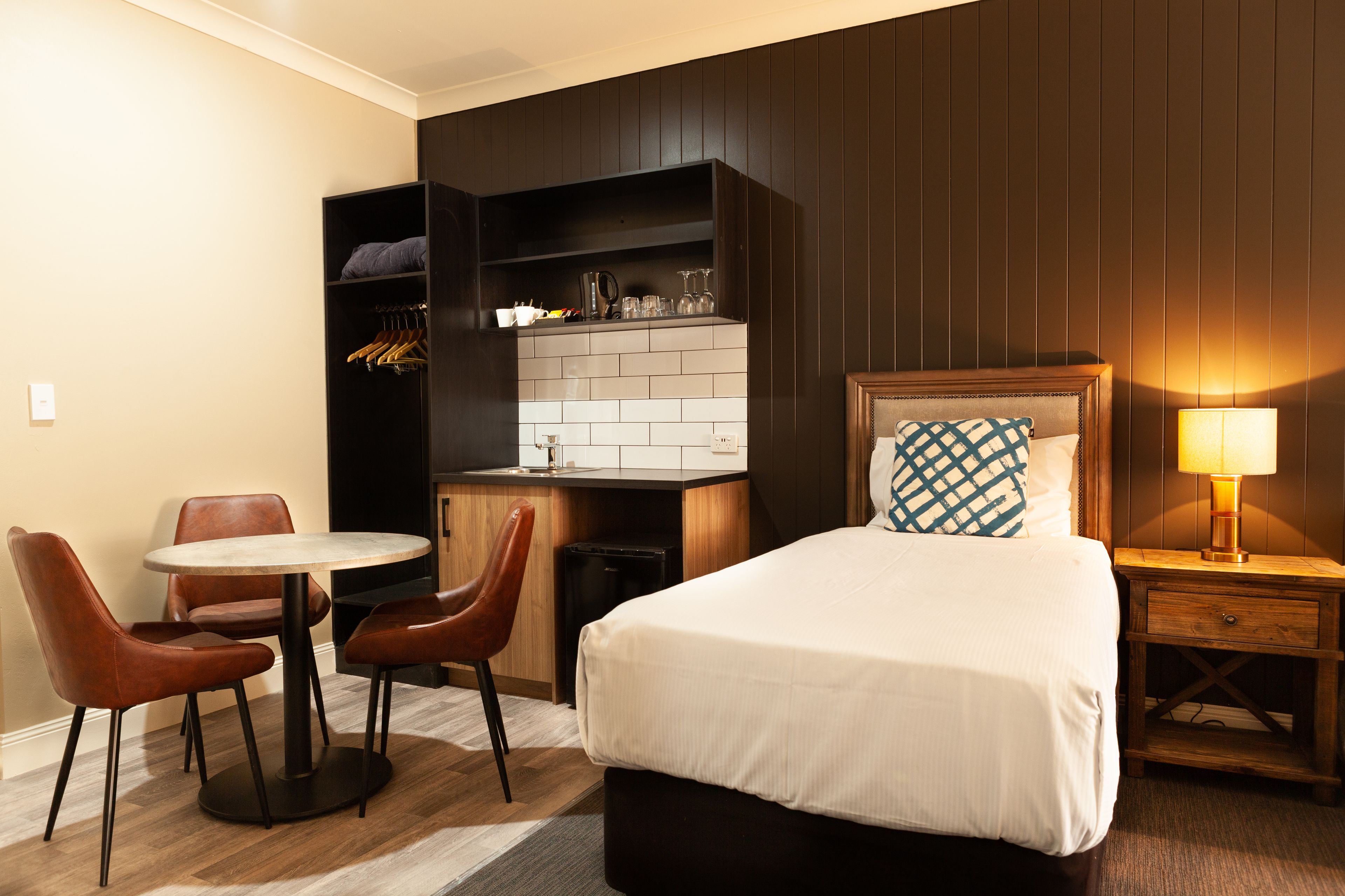 A hotel room with a bed, bedside table, lamp, and a small kitchenette area with a sink and storage.