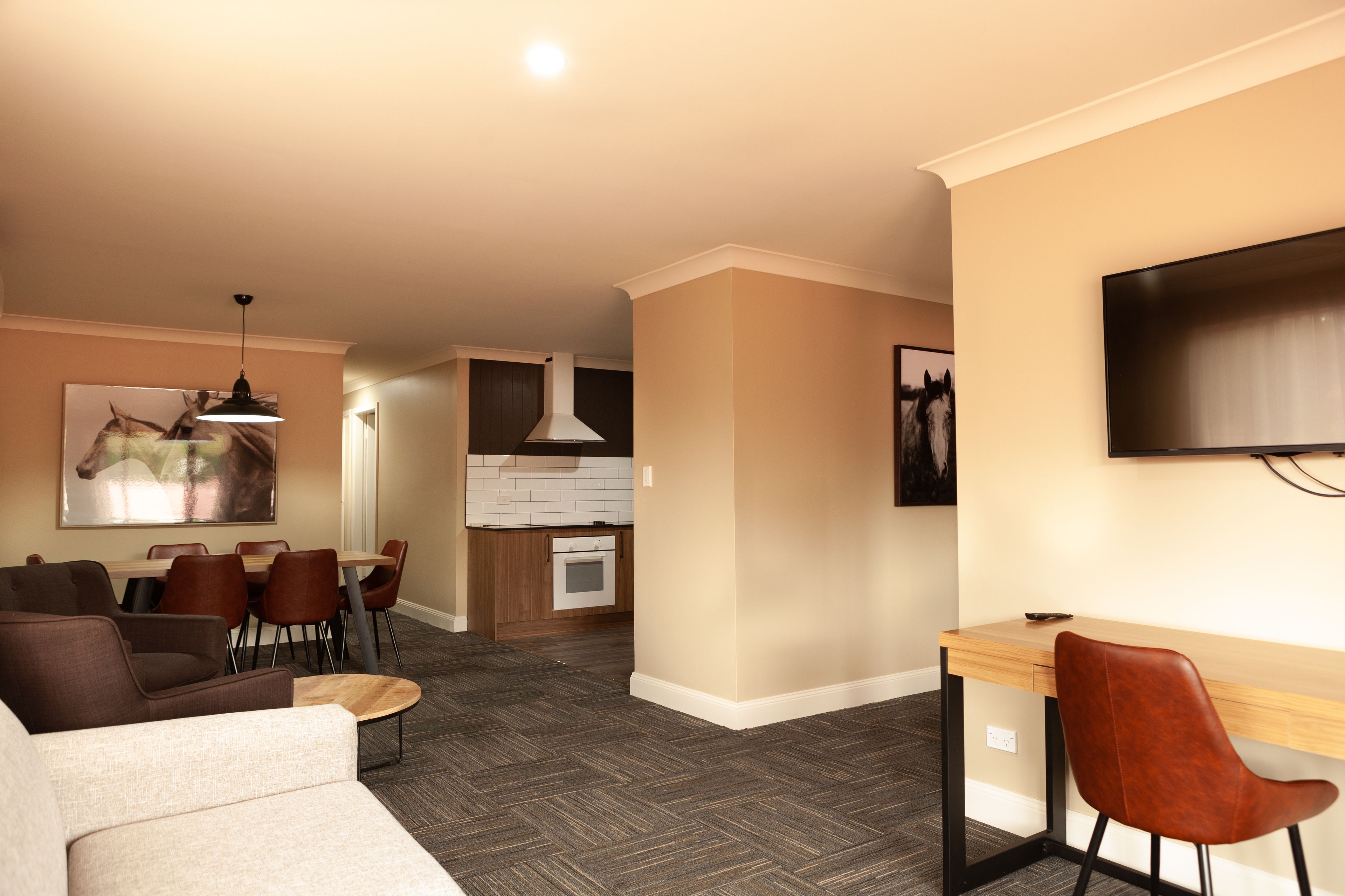 A modern living room with a sofa, dining table, and chairs, a kitchen area with a stove and cabinets, and a flat-screen TV mounted on the wall.